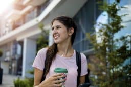 a woman with hearing aids walks past a modern building while holding a reusable coffee cup
